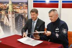 Dwóch mężczyzn podpisuje dokumenty przy stole, jeden w garniturze, drugi w mundurze policji.