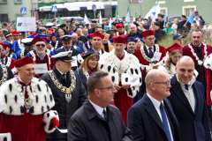 Władze uczelni w czerwonych togach z gronostajowymi pelerynami stoją wśród uczestników plenerowej uroczystości; w tle pozostali goście i zieleń