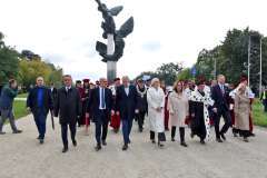 Władze uczelni w czerwonych togach z gronostajowymi pelerynami stoją wśród uczestników plenerowej uroczystości; w tle pozostali goście i zieleń