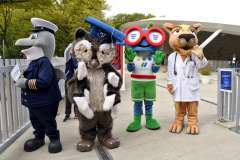 Cztery maskotki – marynarz, sowa, zielona postać w dużych okularach i puma w fartuchu lekarskim – stoją obok siebie przed budynkiem