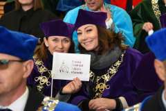 Dwie kobiety w fioletowych togach i biretach siedzą wśród uczestników uroczystości, uśmiechają się do zdjęcia i trzymają małą flagę z logo PUM