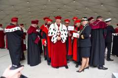 Osoby w czerwonych i czarnych togach akademickich stoją pod zadaszeniem podczas uroczystości plenerowej; w centrum mężczyzna w czerwonej todze 