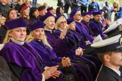 Osoby w fioletowych togach i biretach siedzą w rzędzie na widowni podczas uroczystości; w tle widoczni są inni uczestnicy wydarzenia