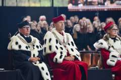 Trzy osoby w togach rektorskich z łańcuchami insygniów siedzą na scenie podczas uroczystości, w tle widoczna publiczność