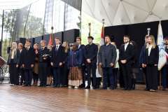 Grupa kilkunastu osób stoi na scenie podczas uroczystości plenerowej; w tle widoczne są poczty sztandarowe