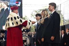 Rektor w czerwonym stroju z gronostajowym kołnierzem pasuje studentkę berłem podczas uroczystości na scenie; obok stoją dwie osoby w garniturach