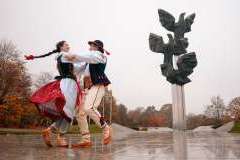 Para w tradycyjnych polskich strojach ludowych tańczy przed pomnikiem trzech orłów na tle jesiennych drzew.