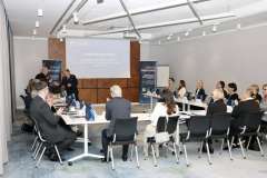 grupa osób siedzi w sali konferencyjnej, na ścianie wisi ekran na którym wyświetla się slajd prezentacji