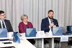 jedna kobieta i dwóch mężczyzn siedzą przy stole, na którym leżą dokumenty. Jeden mężczyzna mówi przez mikrofon