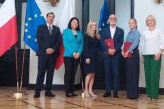 grupa stojących osób w sali, w tle flagi Unii Europejskiej oraz Polski