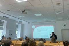 Grupa ludzi siedzi w sali wykładowej i słucha prelegentki. W tle ekran z prezentacją