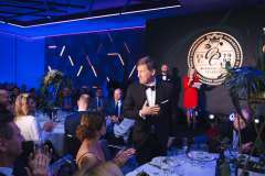 Mężczyzna w smokingu wstaje od stołu i kłania się podczas Gali Biznesu, a publiczność siedząca przy stołach bije brawo