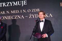 Mężczyzna w smokingu stoi przy mikrofonie na scenie Gali Biznesu i przemawia, trzymając teczkę z dokumentami