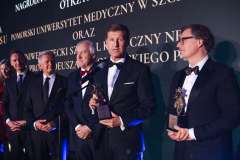 Laureaci w smokingach stoją na scenie podczas Gali Biznesu, trzymając statuetki i pozując do zdjęcia na tle ekranu z nazwą wydarzenia.