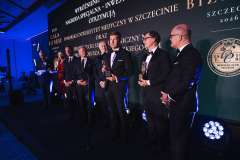 Grupa mężczyzn w smokingach stoi na scenie podczas Gali Biznesu w Szczecinie, trzymając statuetki i rozmawiając po zakończeniu wręczenia nagród