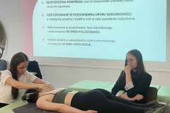 W sali warsztatowej. Na tle ekranu z prezentacją znajdują się trzy kobiety. Dwie prezentują techniki masażu, jedna siedzi i obserwuje