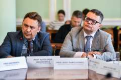 Dwóch mężczyzn siedzących przy stole. Jeden w okularach korekcyjnych, ubrani w garnitury i krawaty