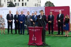 mężczyzna przemawia przy mównicy, w tle stoi grupa osób, ścianka reklamowa z logotypami oraz budynek szpitala