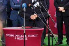 mężczyzna przemawia przy mównicy, w tle stoi grupa mężczyzn oraz ścianka reklamowa z logotypami