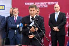 mężczyzna przemawia przy mównicy, w tle stoi grupa osób, ścianka reklamowa z logotypami 