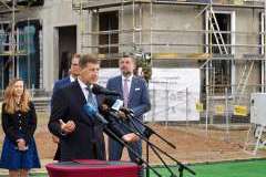 mężczyzna przemawia przy mównicy, w tle stoją inne osoby, za nimi teren budowy