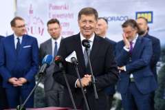 mężczyzna przemawia przy mównicy, w tle stoi grupa mężczyzn oraz ścianka reklamowa z logotypami