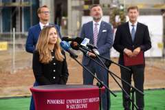 kobieta przemawia przy mównicy, w tle stoją trzej mężczyźni