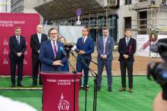 mężczyzna przemawia przy mównicy, w tle stoi grupa osób, ścianka reklamowa z logotypami, teren budowy