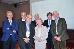 Grupa siedmiu uczestników konferencji stoi razem i pozuje do zdjęcia; wszyscy mają identyfikatory, a w tle widać ekran projekcyjny