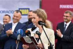 kobieta przemawia przy mównicy, w tle stoi grupa osób, ścianka reklamowa z logotypami 