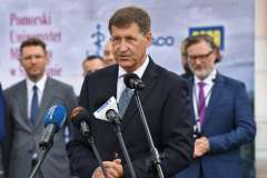 mężczyzna przemawia przy mównicy, w tle stoi grupa mężczyzn oraz ścianka reklamowa z logotypami