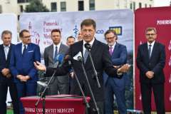 mężczyzna przemawia przy mównicy, w tle stoi grupa mężczyzn oraz ścianka reklamowa z logotypami