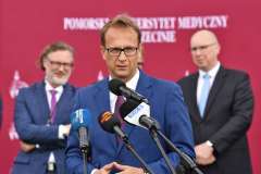 mężczyzna przemawia przy mównicy, w tle stoi trzech mężczyzn, ścianka reklamowa z logotypami