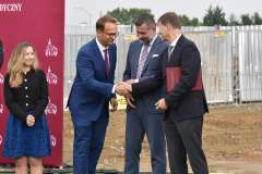 stoi kobieta i trzech mężczyzn, dwóch z nich podaje sobie rękę, w tle teren budowy