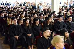 Absolwenci w czarnych togach i biretach siedzą w auli podczas uroczystości; w tle widoczna jest publiczność wypełniająca rzędy miejsc