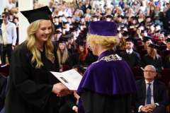 Uśmiechnięta absolwentka w czarnej todze i birecie odbiera dyplom od przedstawicielki władz uczelni w fioletowej todze podczas uroczystości