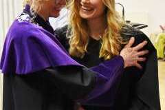 Przedstawicielka władz uczelni w fioletowej todze obejmuje uśmiechniętą absolwentkę w czarnej todze i birecie; absolwentka trzyma w dłoni małą torbę 