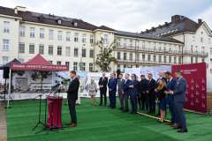 mężczyzna przemawia przy mównicy, w tle stoi grupa osób, ścianka reklamowa z logotypami oraz budynek szpitala