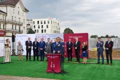 mężczyzna przemawia przy mównicy, w tle stoi grupa osób, ścianka reklamowa z logotypami oraz budynek szpitala