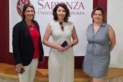 Trzy kobiety stojące przed tablicą z logo Sapienza University of Rome. Wszystkie uśmiechają się do kamery. 