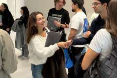 grupa młodych osób stojących na korytarzu, młoda kobieta podaje rękę młodemu mężczyźnie 