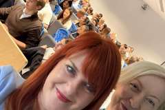 dwie uśmiechnięte kobiety , w tle młodzi ludzie siedzący w sali wykładowej