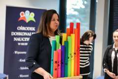 Kobieta podczas prezentacji trzyma zestaw kolorowych rur o różnej wysokości; w tle stoją dwie osoby oraz baner Ośrodka Doskonalenia Nauczycieli