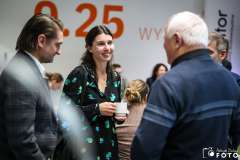 Kilka osób rozmawia podczas przerwy w wydarzeniu; kobieta w czarnej sukience z zielonym wzorem trzyma filiżankę i uśmiecha się do rozmówców