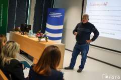 Mężczyzna prowadzi prezentację w sali szkoleniowej, stojąc obok ekranu z wyświetlonym slajdem; przed nim siedzą uczestnicy, a na biurku stoją kwiaty 