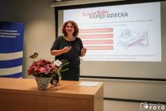 Kobieta prowadzi prezentację w sali szkoleniowej, stojąc obok ekranu z wyświetlonym slajdem a na biurku stoją kwiaty i materiały szkoleniowe