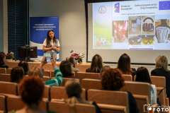 Kobieta prowadzi prezentację w sali szkoleniowej, stojąc obok ekranu z wyświetlonym slajdem; przed nią siedzą uczestnicy