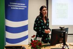 Kobieta prowadzi prezentację w sali szkoleniowej, stojąc obok ekranu z wyświetlonym slajdem a na biurku stoją kwiaty i materiały szkoleniowe