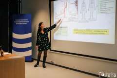 Kobieta prowadzi prezentację w sali szkoleniowej, stojąc obok ekranu z wyświetlonym slajdem wskazuje lewą ręką na slajd