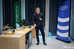 Mężczyzna prowadzi wystąpienie w sali szkoleniowej, stojąc obok biurka z komputerem i materiałami; w tle widoczne są banery informacyjne 